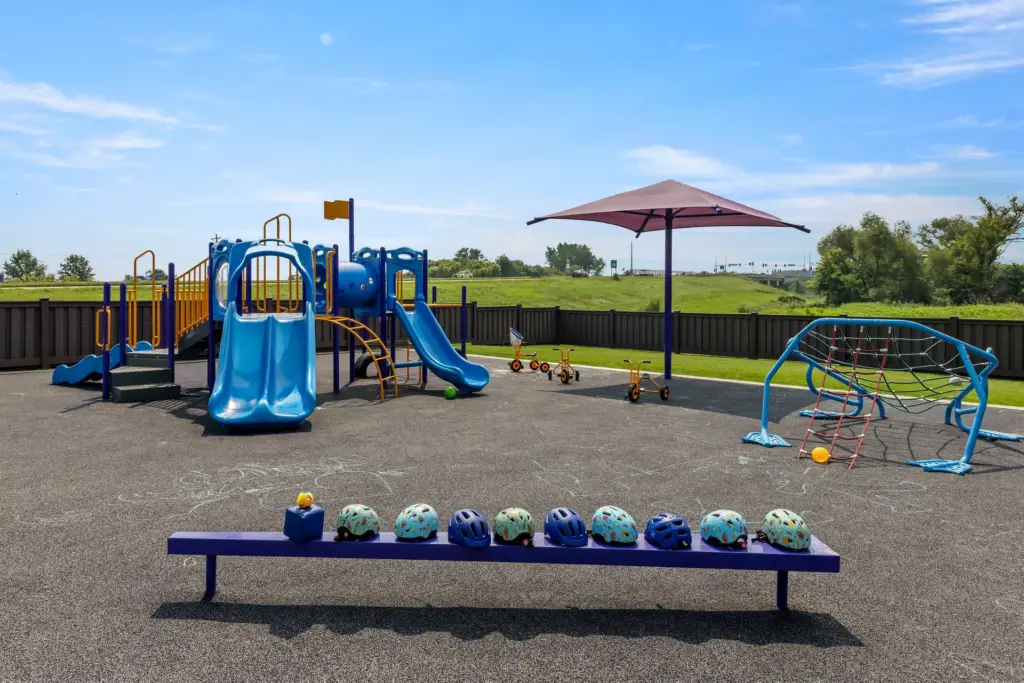 The playground at the Rochester facility. There’s a bench with bike helmets, an umbrella, tricycles, and a large play structure with slides and spots to climb.
