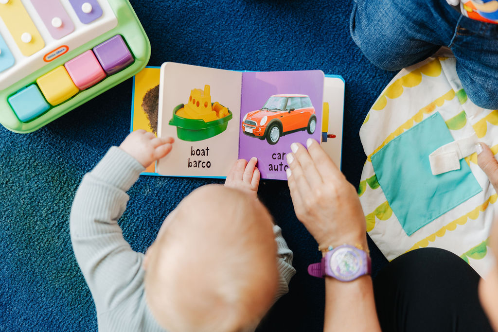 A young child reading from a board book with the help of a teacher.